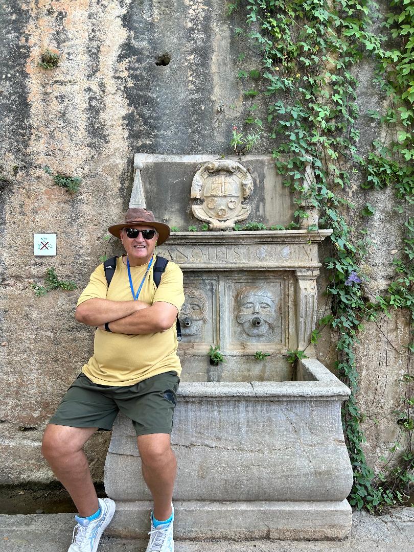 A man wearing a wide-brimmed hat and sunglasses sits on a concrete statue with vines around it.