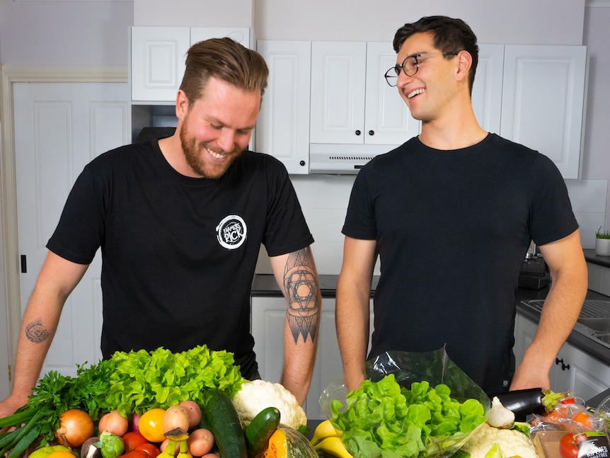 Two young men wearing black t-shirts stand together smiling fresh produce on a bench.