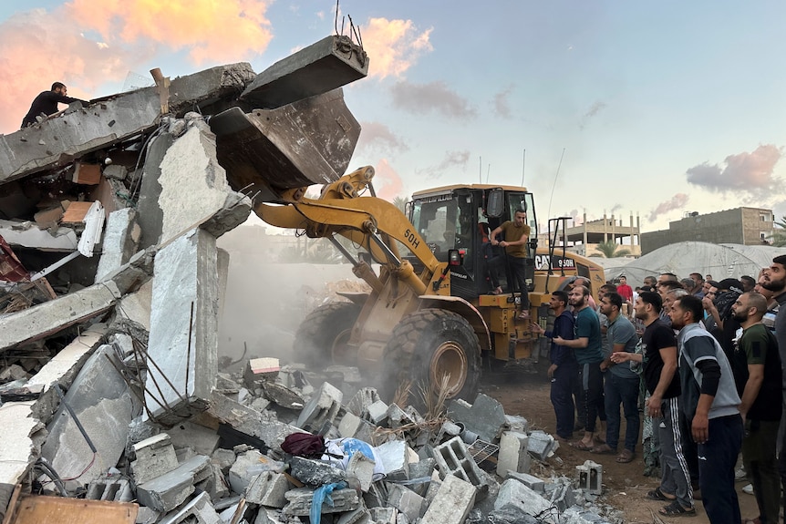A truck lifts the debris of a building as people watch on 