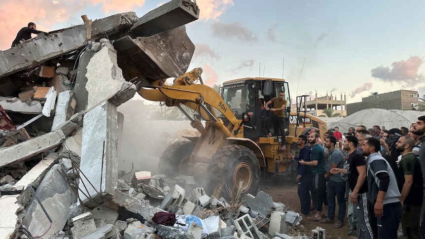 A truck lifts the debris of a building as people watch on 