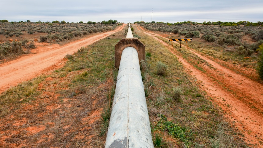 A pipeline rises above ground surrounded by red dirt and grass and goes off into the distance