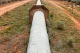 A pipeline rises above ground surrounded by red dirt and grass and goes off into the distance