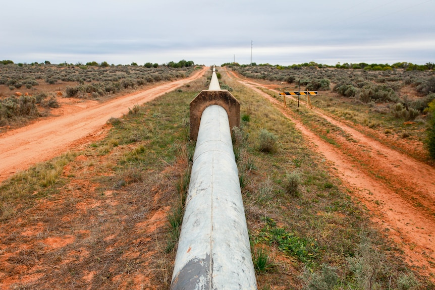 A pipeline rises above ground surrounded by red dirt and grass and goes off into the distance