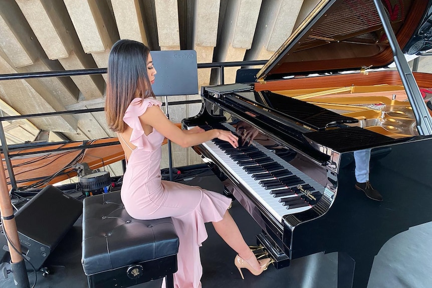 A young woman wearing a pink dress plays a grand piano.