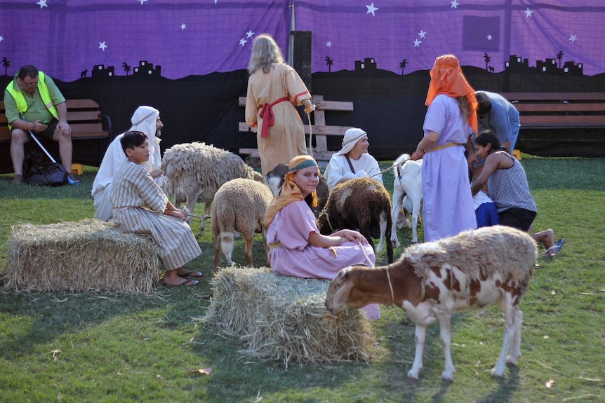 A group of children and young adults dressed as shepherds sit on hay bales with sheep in the middle.