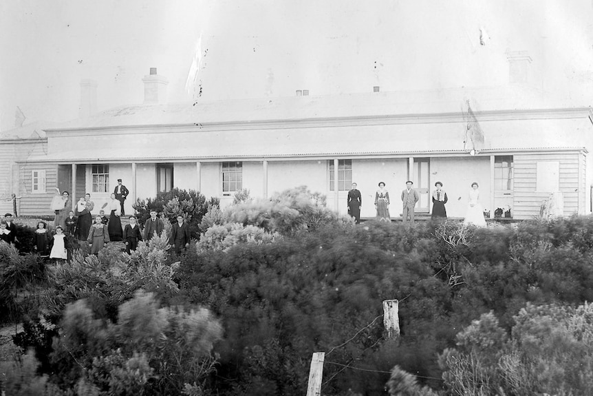 Men, women and children line a wide verandah at the front of a weatherboard cottage.