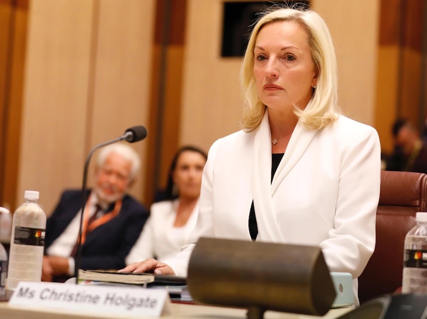 Christine Holgate standing at a senate committee hearing, she is pictured with a white blazer