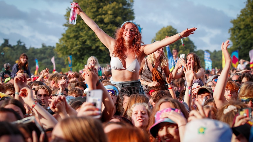 A girl sitting on someone's shoulders outstretches her arms and grins at a packed outdoor mosh pit 