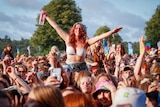 A girl sitting on someone's shoulders outstretches her arms and grins at a packed outdoor mosh pit 