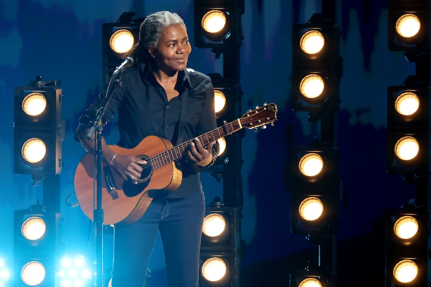 Tracy Chapman wearing dark clothing, hair tied back, holding a guitar, stage lights behind her
