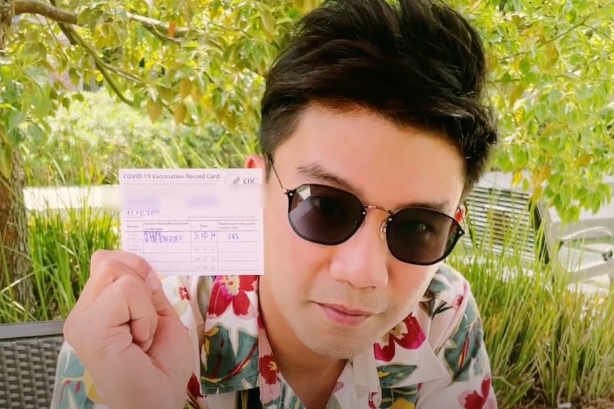 A young Thai man in black sunglasses holds a vaccination card 