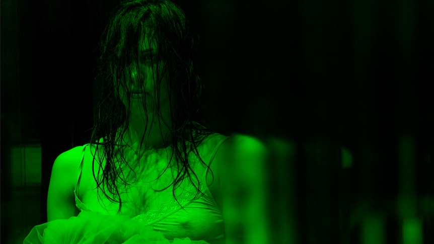 Woman with wet dark brown long hair over face in greenish light and darkened theatre, wearing white singlet and holding tulle.