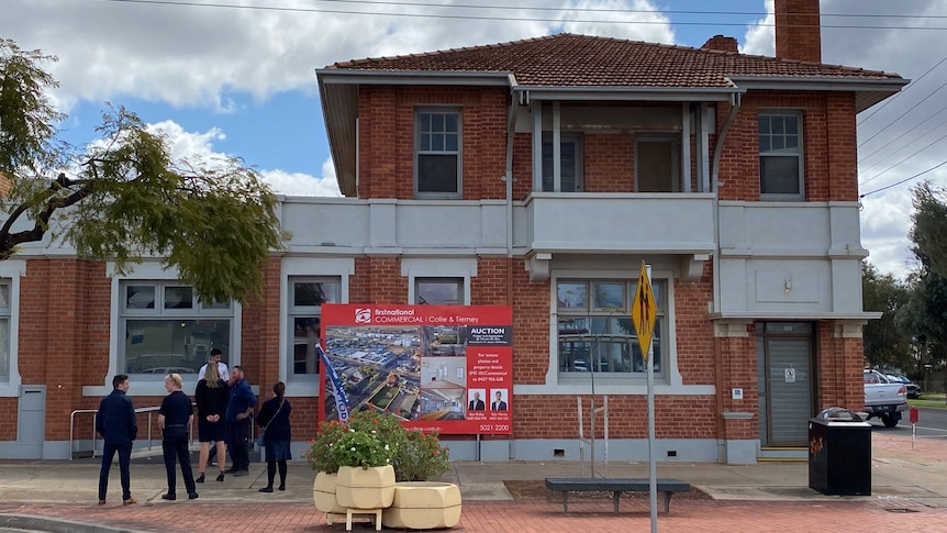 A small crowd gathers outside of a brick, two-storey bank building with a "for sale" sign across the front.