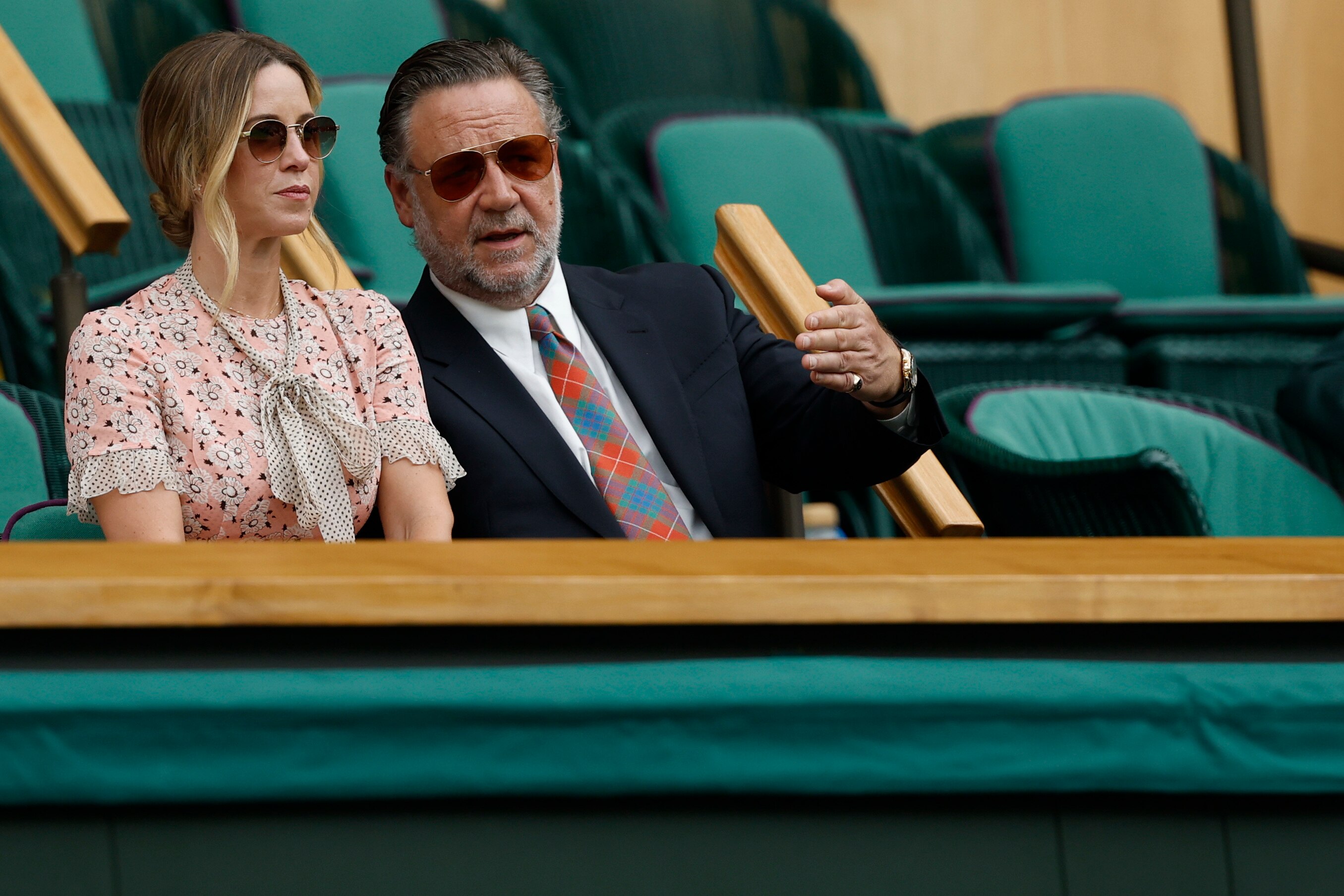 Russell Crowe speaks to his partner in a floral dress while he wears a dark suit and colourful tie 