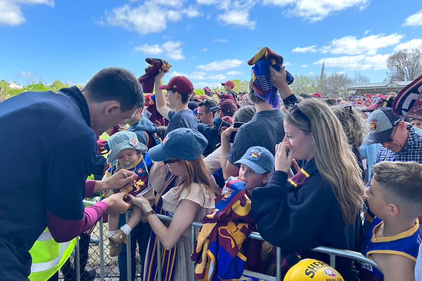A Lions player signs autographs as crowds of fans, many with Lions scarves and hats, stand against a barrier.