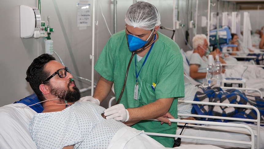 A man reclines in a hospital bed while another man in green scrubs stands by his side