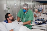 A man reclines in a hospital bed while another man in green scrubs stands by his side