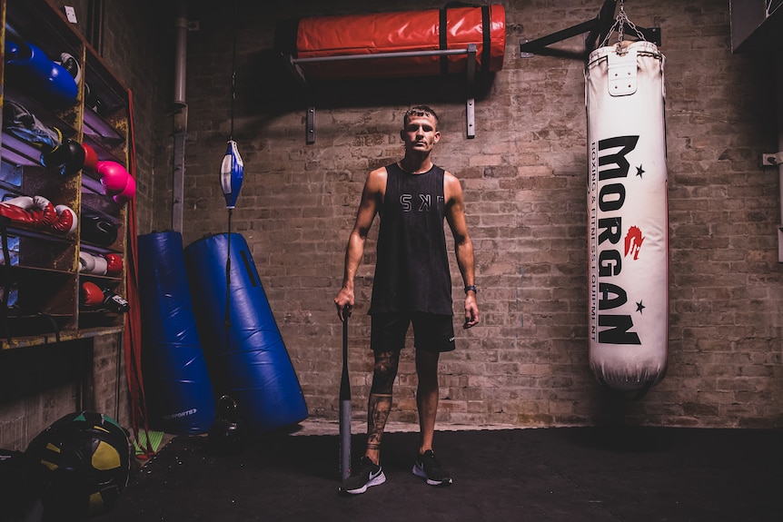 A man in exercise gear in a room holding a baseball bat, standing next to light and heavy bags and other gear. 