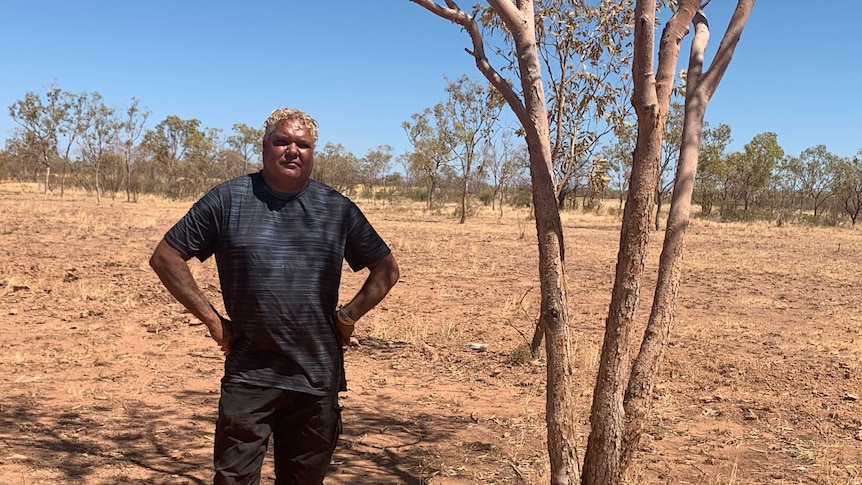 An Indigenous man with grey hair stands in sparse bushland with his hands on his hips.