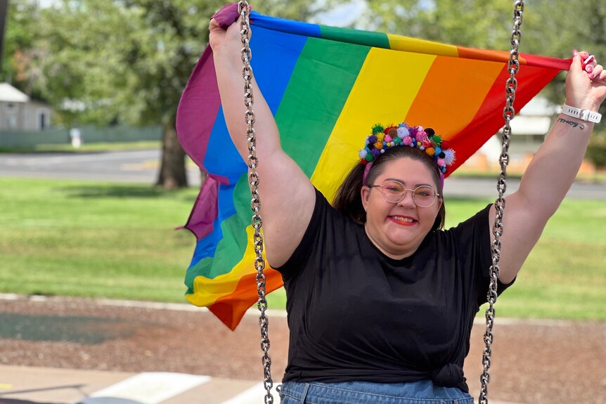 A woman sits on a swing at a park, with a rainbow flag streaming out behind her