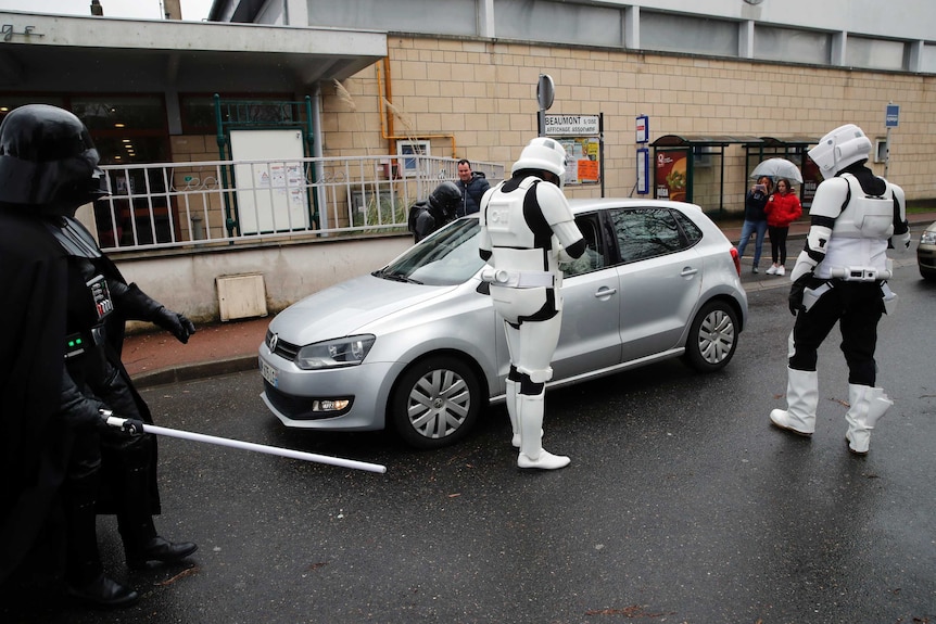 Two men in Star Wars stormtrooper gear stand next to car while a man in a Darth Vader suit watches.