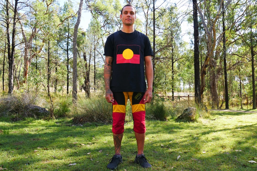 An Indigenous man in a black shirt with the Aboriginal flag stands and looks at the camera.