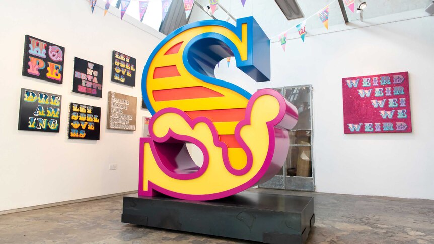 A gallery room with street-art style paintings on the wall and a large, brightly-coloured letter 'S' sculpture at the centre.