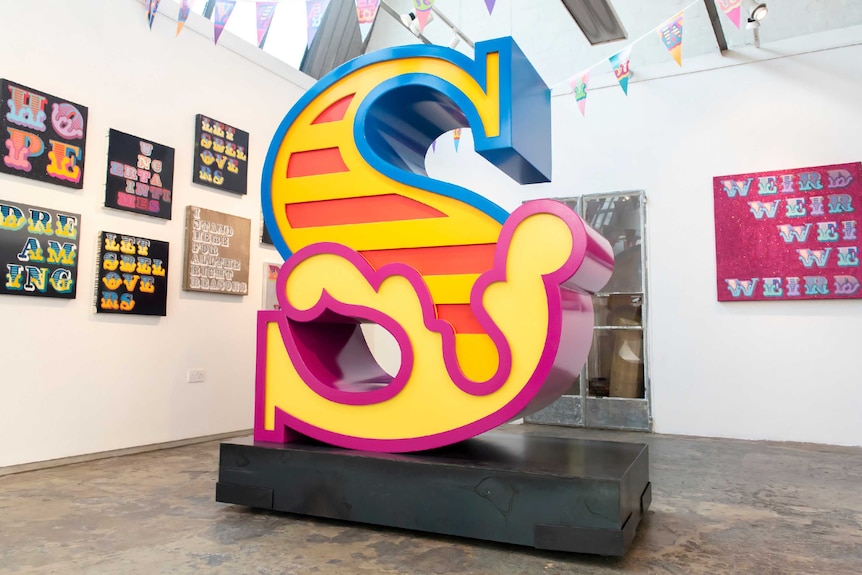 A gallery room with street-art style paintings on the wall and a large, brightly-coloured letter 'S' sculpture at the centre.