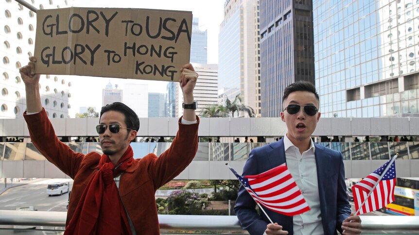 You look at two men in a dense Hong Kong district, one holds a cardboard sign and the other holds two US flags.