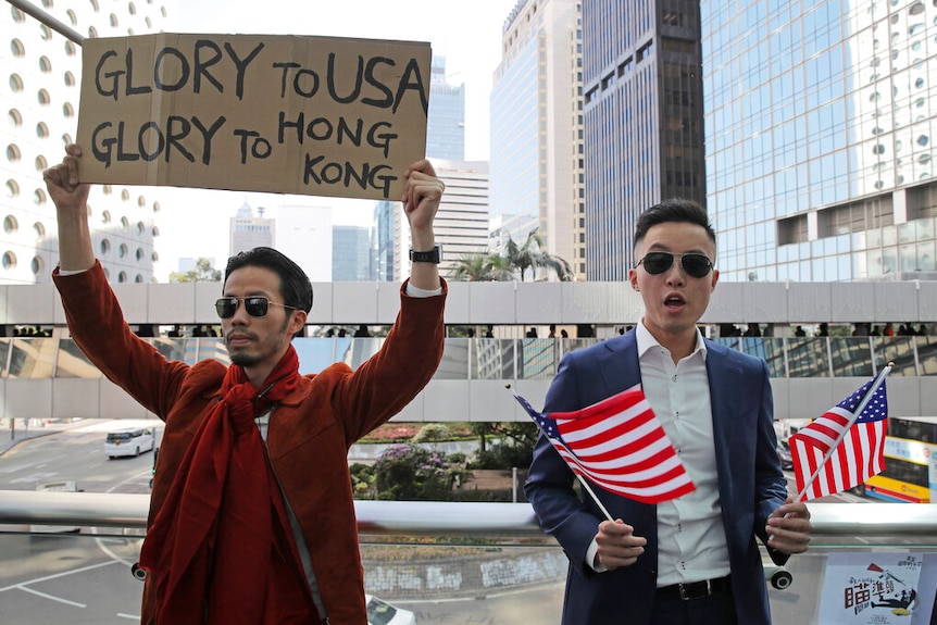 You look at two men in a dense Hong Kong district, one holds a cardboard sign and the other holds two US flags.