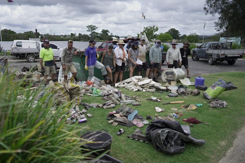 Multiple people standing in front of an empty skip with piles of organised rubbish on the grass in front of them. 