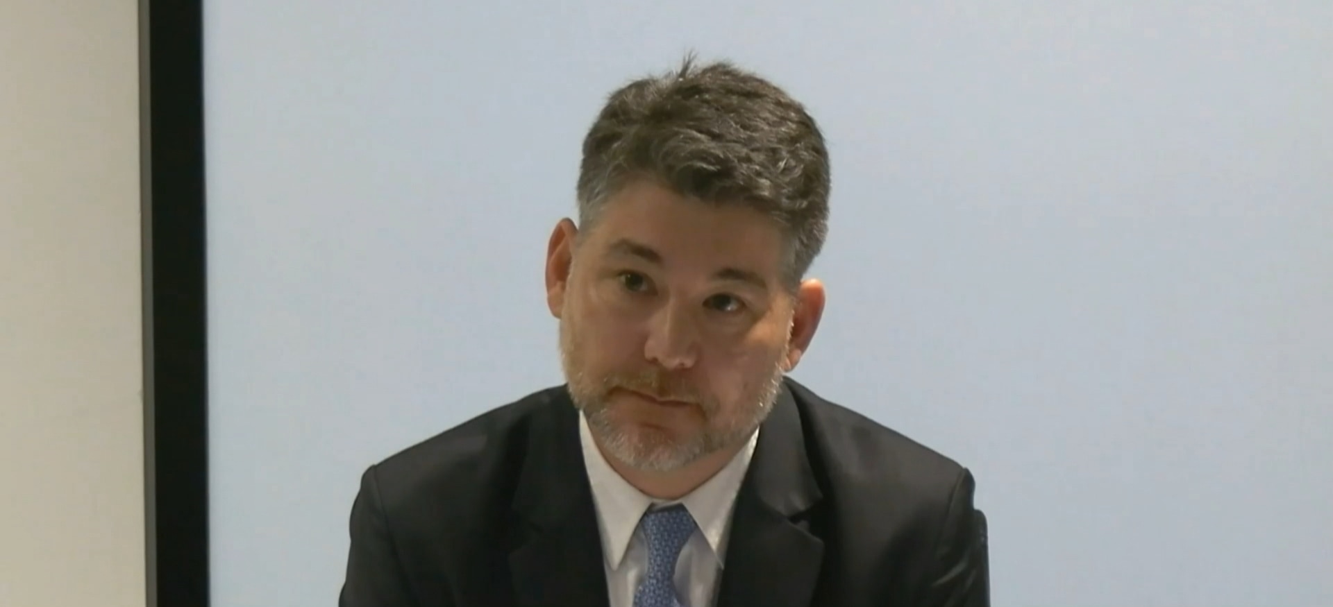 Mr Fang wearing a black suit and blue tie sitting in a chair looking serious.