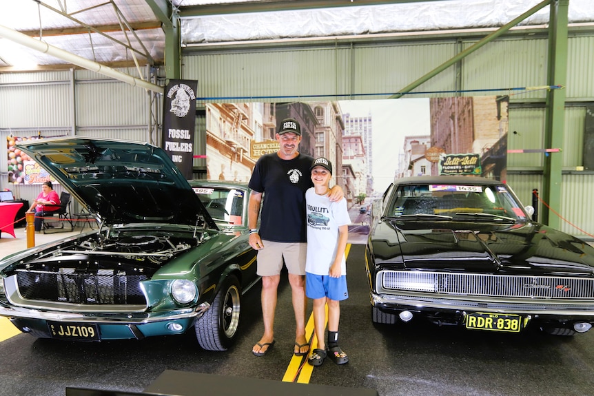 A man wearing a black shirt and boy wearing white shirt stand between a green Ford Mustang GT and black Dodge Charger.