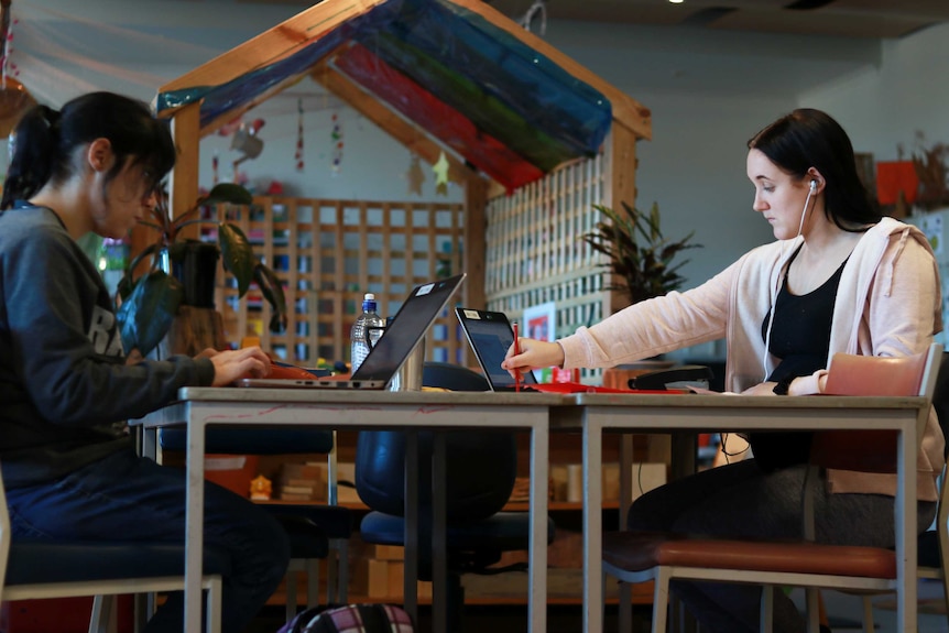 Two young women do schoolwork on laptops with a classroom nursery in background