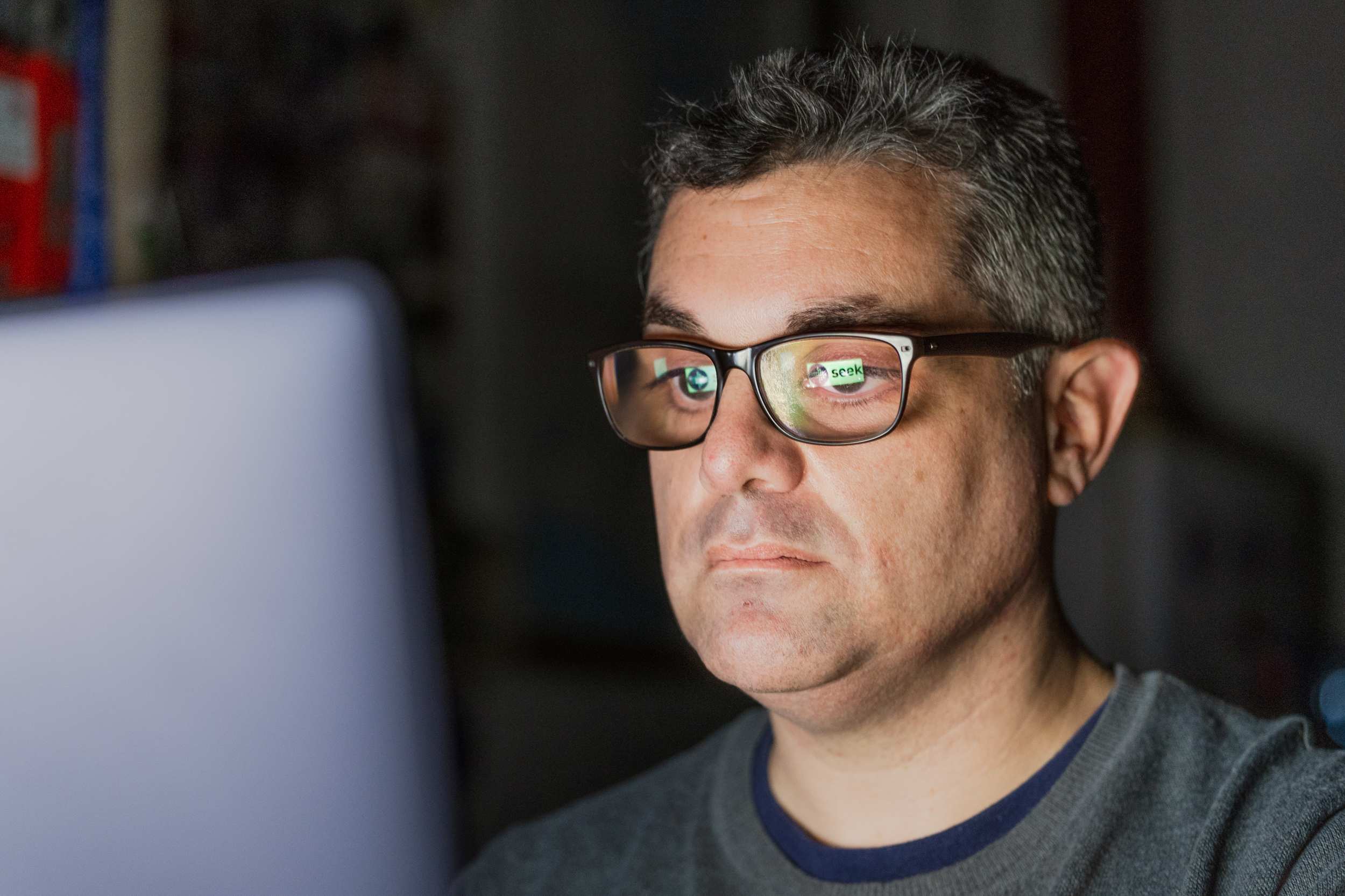 A man with glasses and a grey jumper sits at a laptop. A Seek website logo is reflected in his glasses.