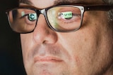 A man with glasses and a grey jumper sits at a laptop. A Seek website logo is reflected in his glasses.