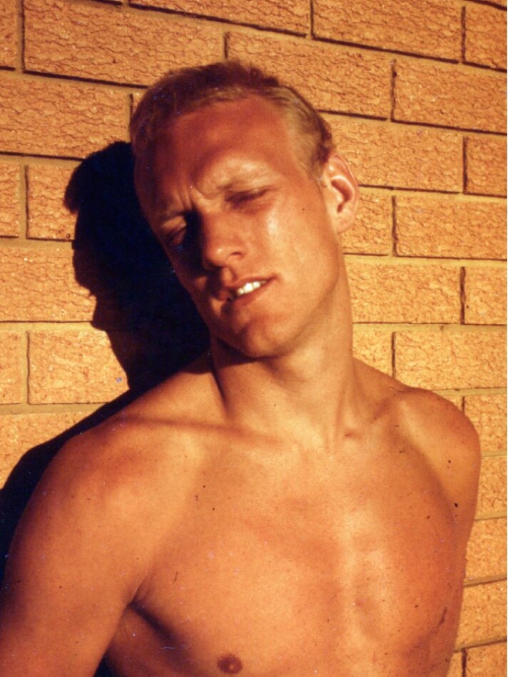 Young man with short, light-coloured hair leaning against a wall, shirtless, the sunlight glow lighting him up 