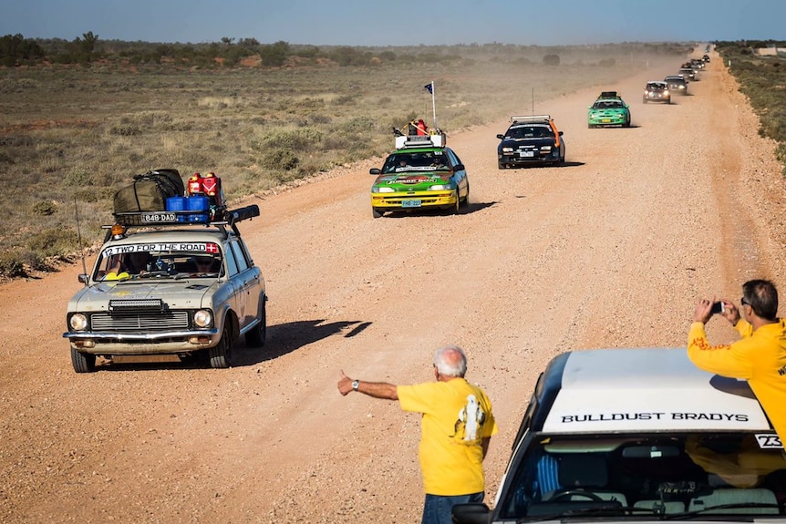A line of cars travelling on a dirt road, two men in the foreground, one giving a thumbs up.
