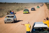 A line of cars travelling on a dirt road. Two men stand in the foreground, one giving a thumbs up.