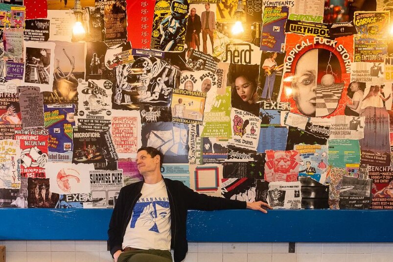 A man sits on a bench against a wall, his arm resting on the bench, many music posters cover the wall.