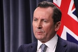 WA Premier Mark McGowan wearing a dark blue suit and tie in front of blue curtains and an Australian flag.