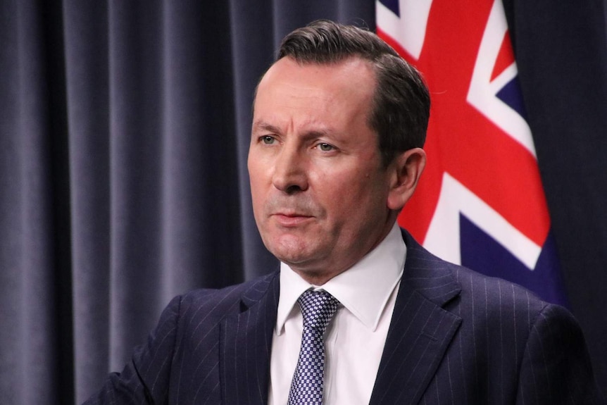 WA Premier Mark McGowan wearing a dark blue suit and tie in front of blue curtains and an Australian flag.