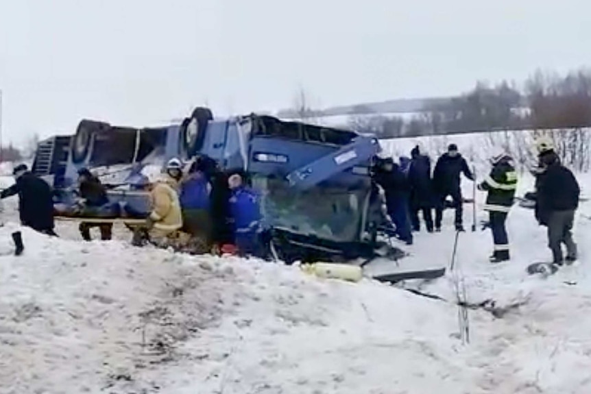 Emergency workers carry an injured bus passenger on a wooden board away from an overturned bus in the snow.