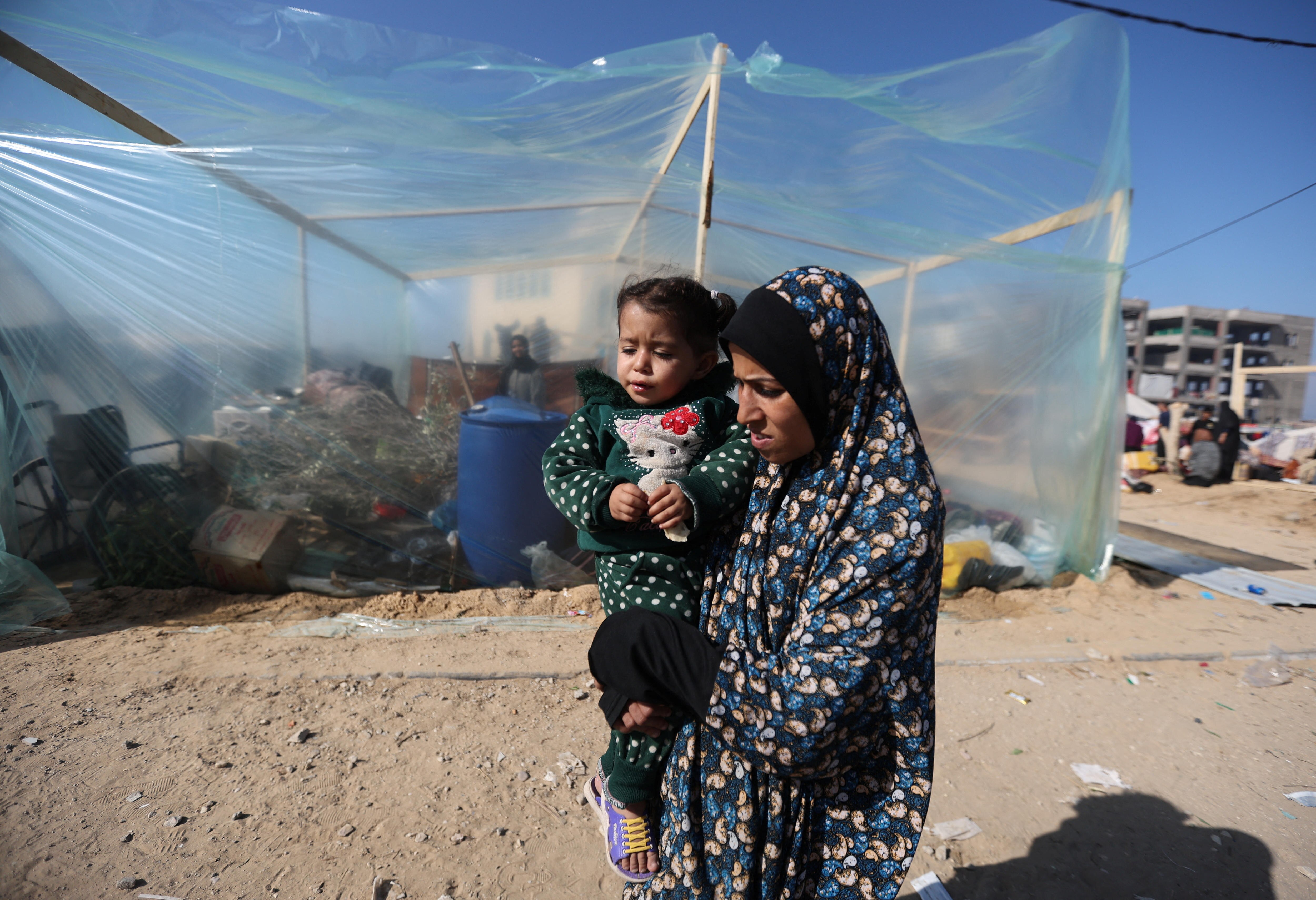 A woman holds a child and stands in front of a plastic tent