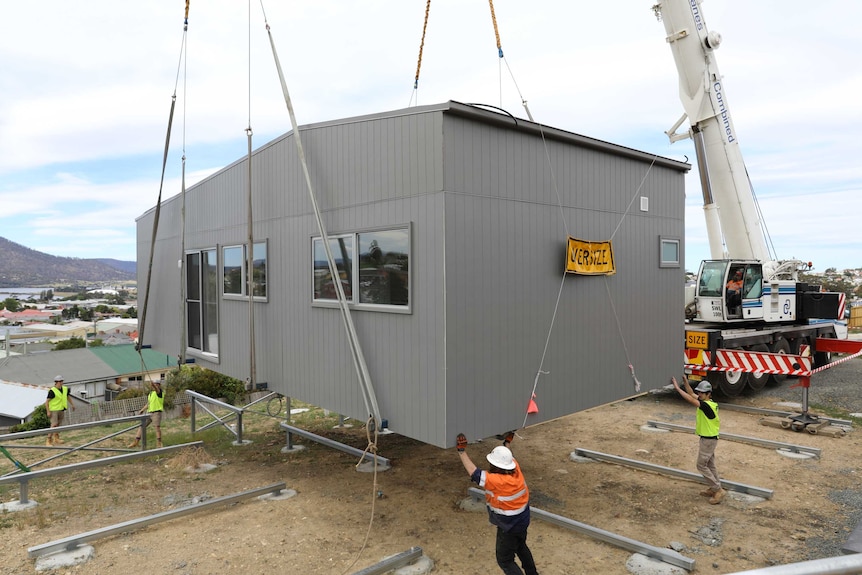 A crane lowers a Supported Affordable Accommodation unit into place at Glenorchy as workmen guide it into place.