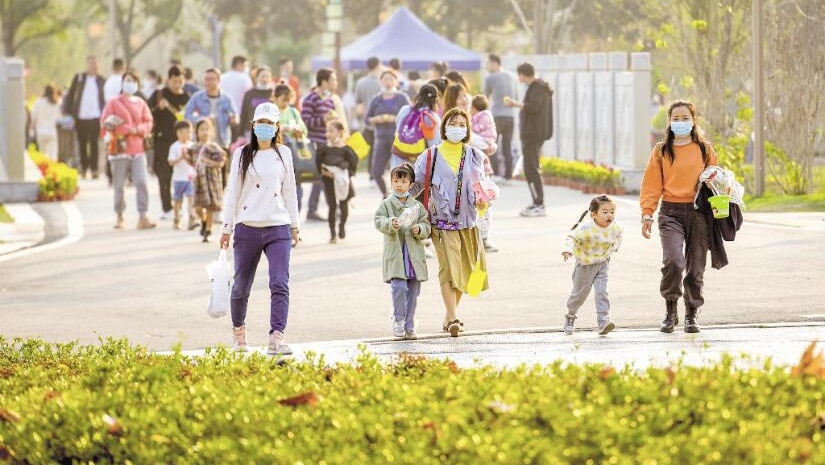 Three women wearing face masks and two girls walk together through a sunny path towards a green plant display.