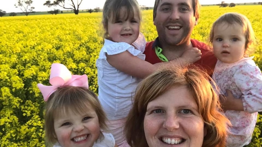 A woman and man holding three young girls smile for a selfie in a yellow field.