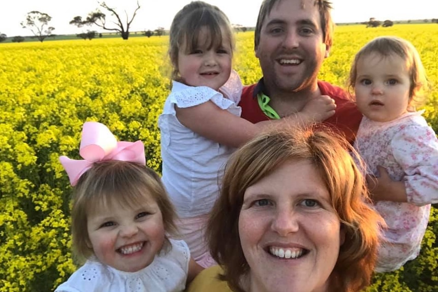 A woman and man holding three young girls smile for a selfie in a yellow field.