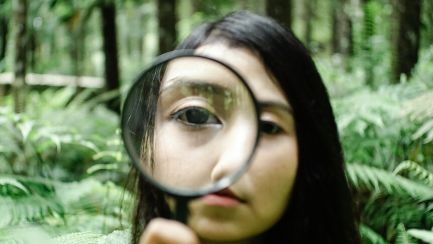 A woman in a forest looking through a magnifying glass held up to her right eye.