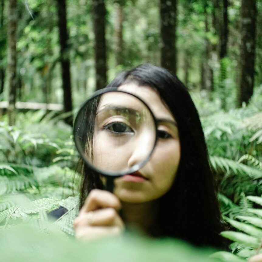 A woman in a forest looking through a magnifying glass held up to her right eye.